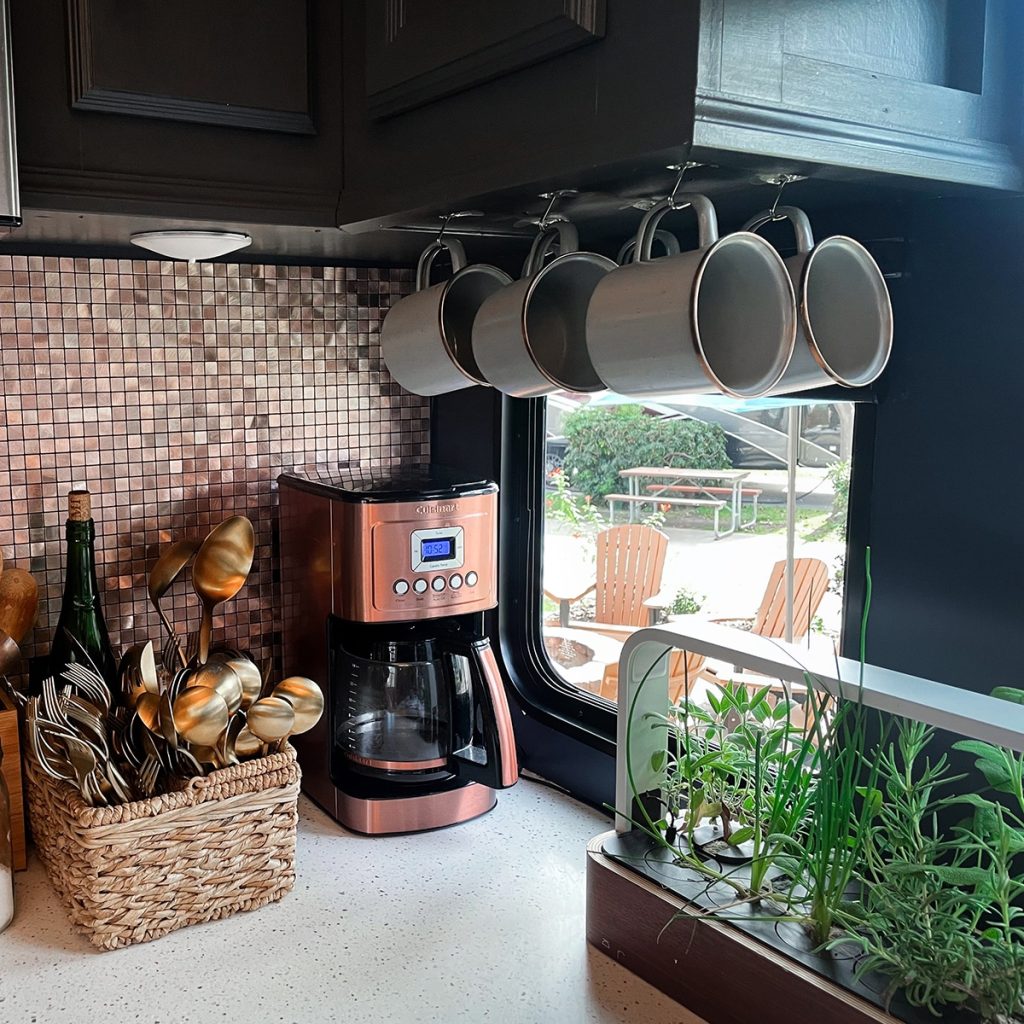 A kitchen corner with a copper coffee maker, hanging mugs, a basket of utensils, and a window herb garden