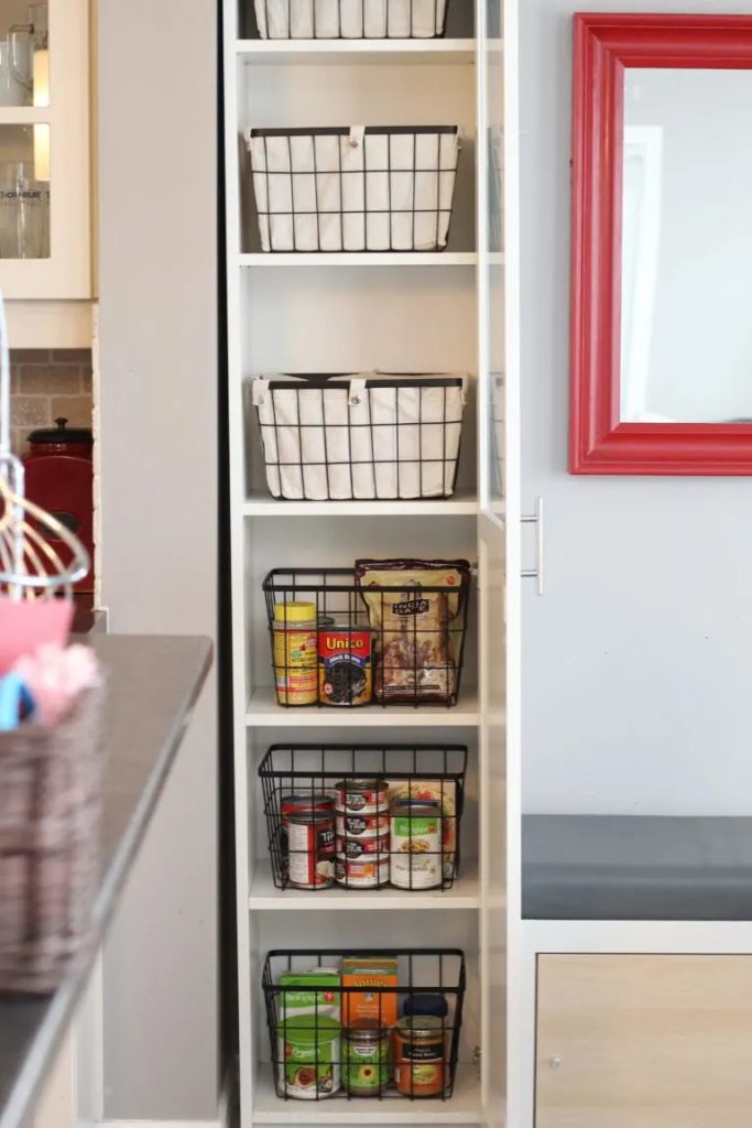 A tall bookcase pantry with shelves organized with white and black wire baskets containing food items and cooking ingredients.