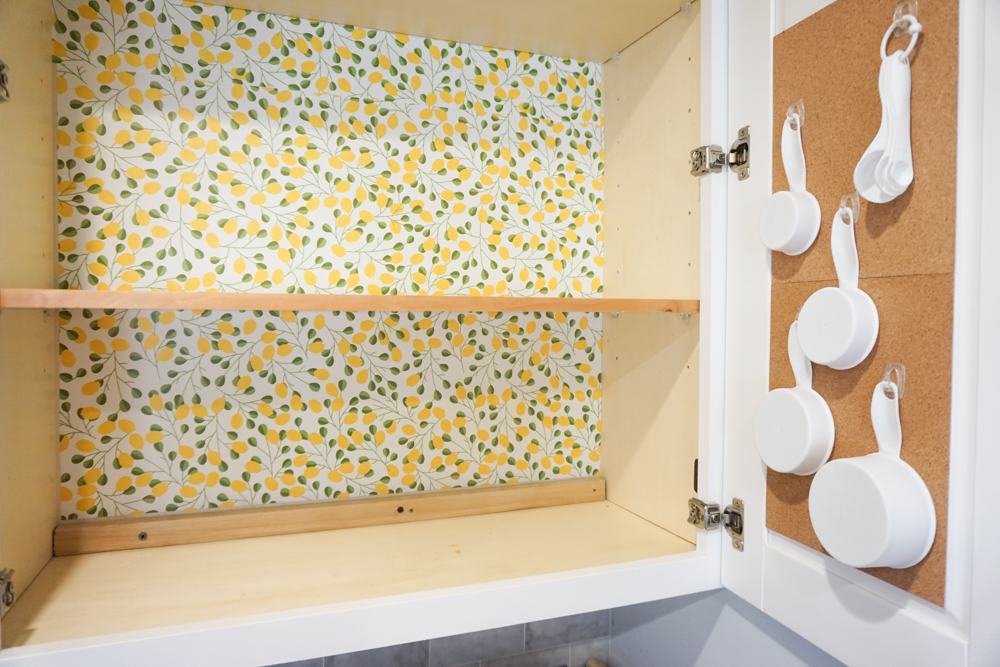 An open cabinet with yellow and green floral wallpaper, wooden shelves, and a cork-lined door holding several hanging white measuring cups