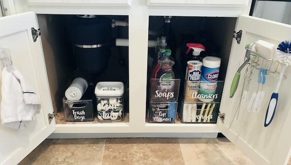 Under-sink storage in a kitchen cabinet with labeled containers for trash bags, soaps, cleansers, and sponges