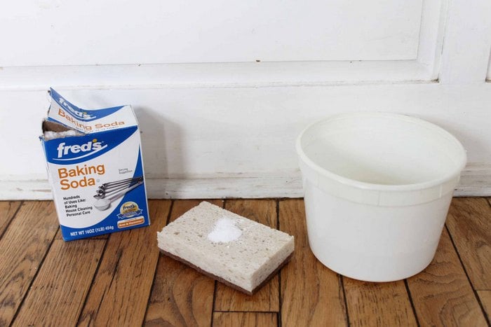 a white bucket, a cleaning sponge with a sprinkle of baking soda on it, and a box of Fred's Baking Soda on a hardwood floor