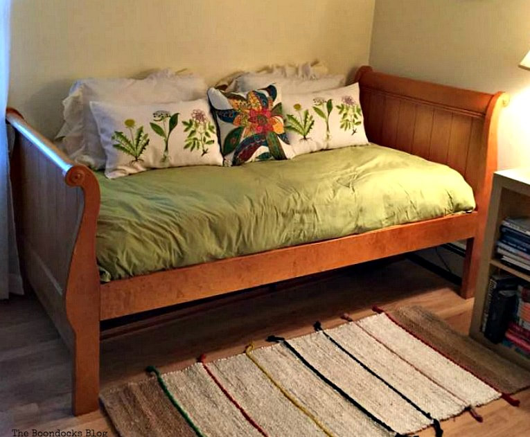 A wooden daybed with green bedding and decorative pillows sits in a  corner with a striped rug
