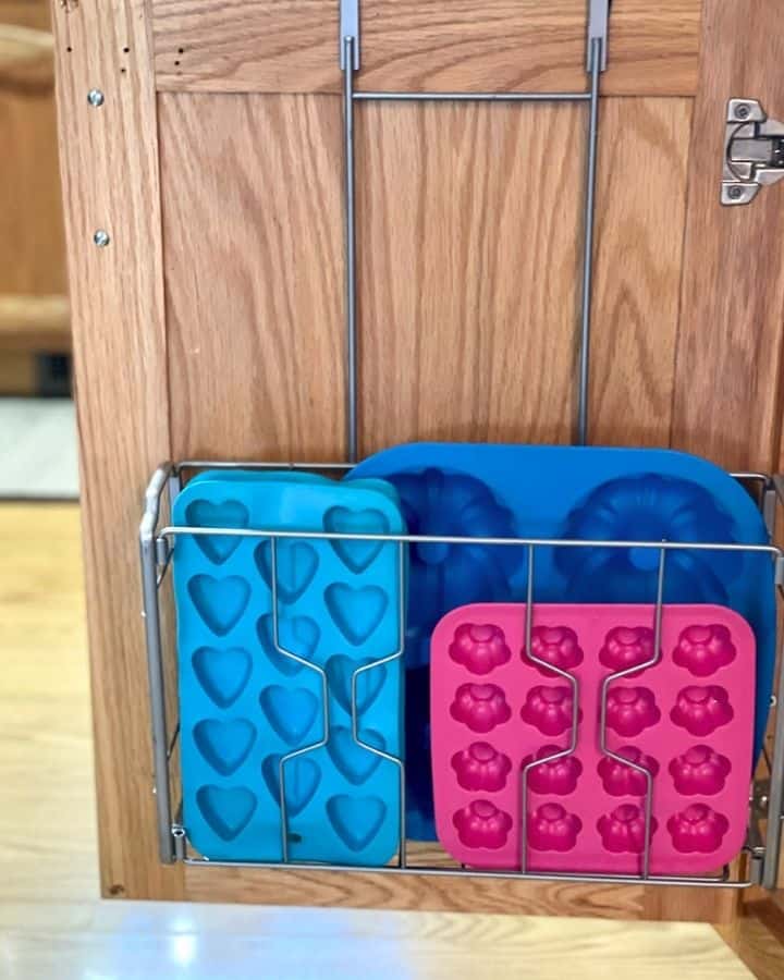 An over-the-door storage rack attached to a wooden cabinet door, holding blue and pink silicone baking molds