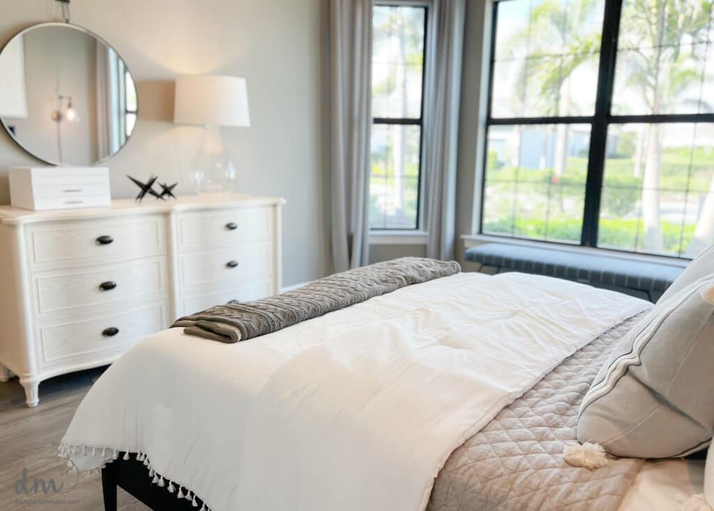 A beautiful calm bedroom with a large window open to a neutral bed with a white dresser