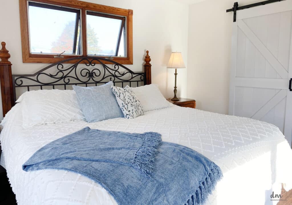 A white bed with a blue blanket and 2 throw pillows next to a lamp on a night stand