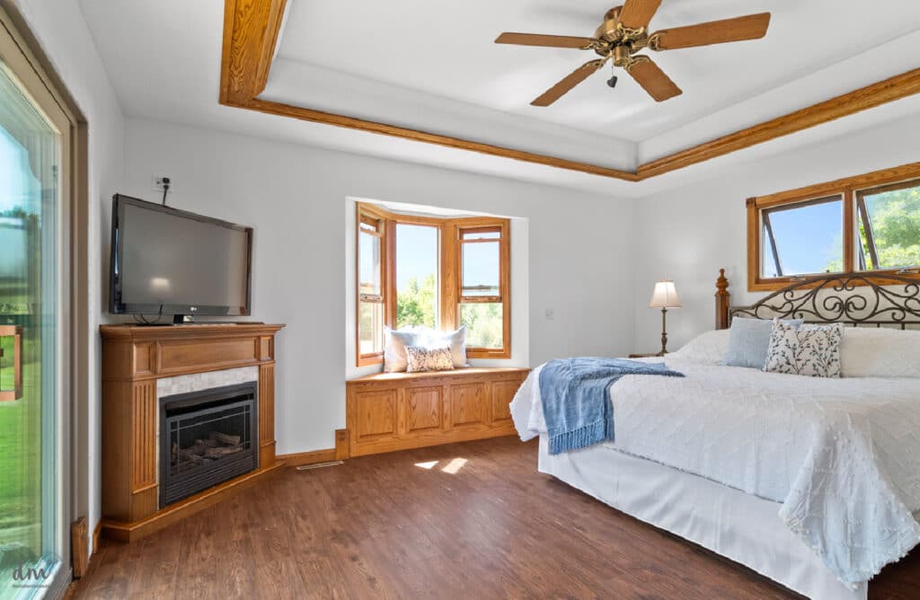 a calm neat bedroom with a white bed next to a window seat and fire place with a tv above
