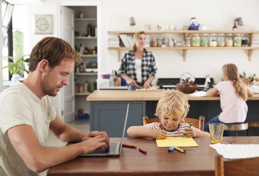 A father works on his laptop while his son colors, and the mother prepares food in the kitchen