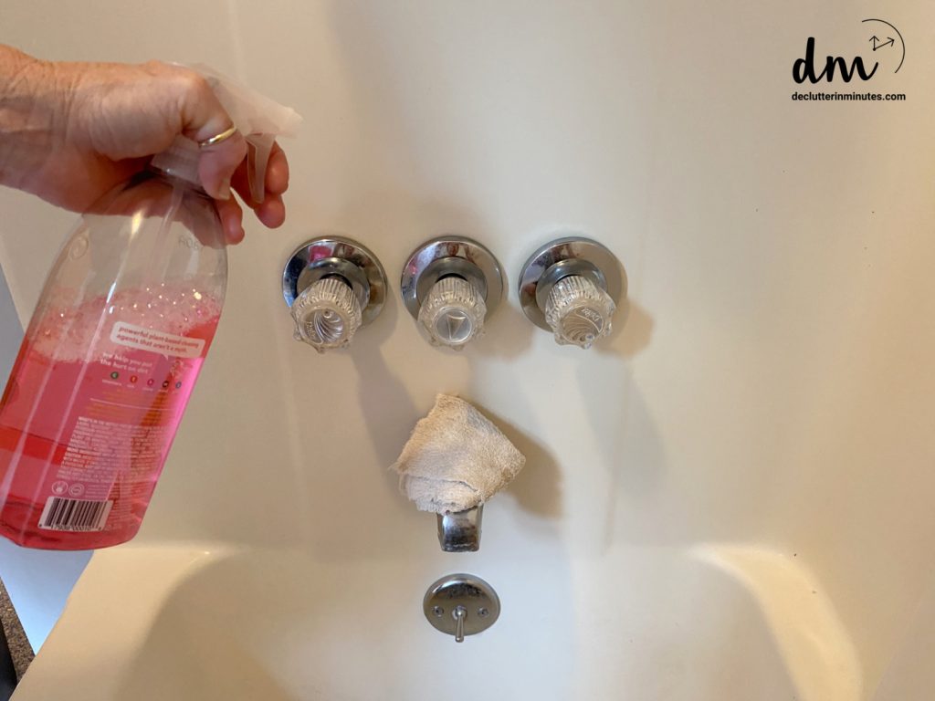 hands holding a bottle of cleaner spraying the faucet in a bathtub