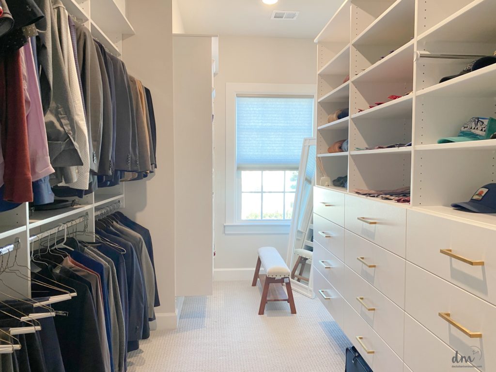 A walk in closet with white shelving and suits hanging down the left side