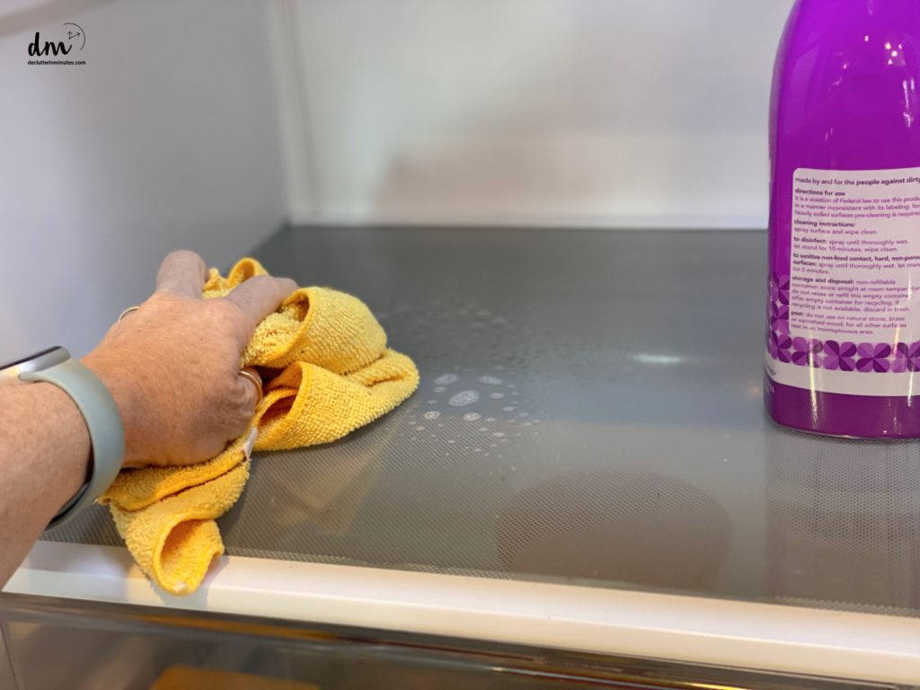 a hand with a yellow cloth cleaning a fridge shelf