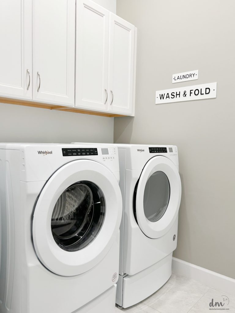 A clean and modern laundry room with a white Whirlpool washer and dryer, overhead cabinets, and a "Wash & Fold" sign on the wall