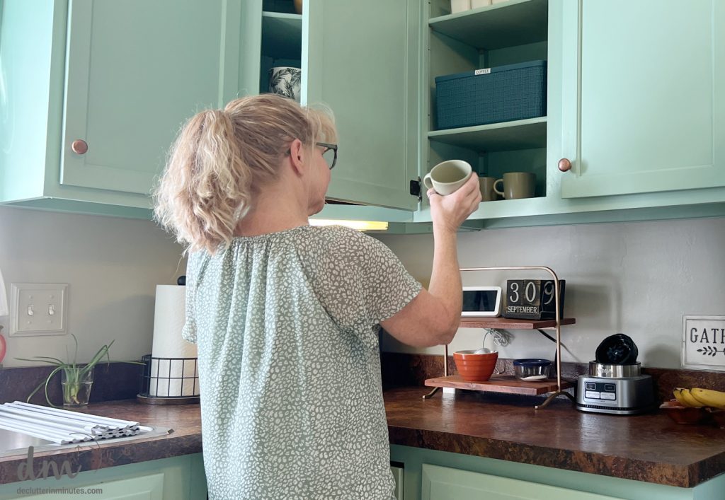 a woman putting coffee mug away in a green cabinet of a kitchen