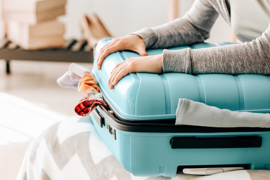 a woman trying to close a teal suitcase that is overflowing with clothes
