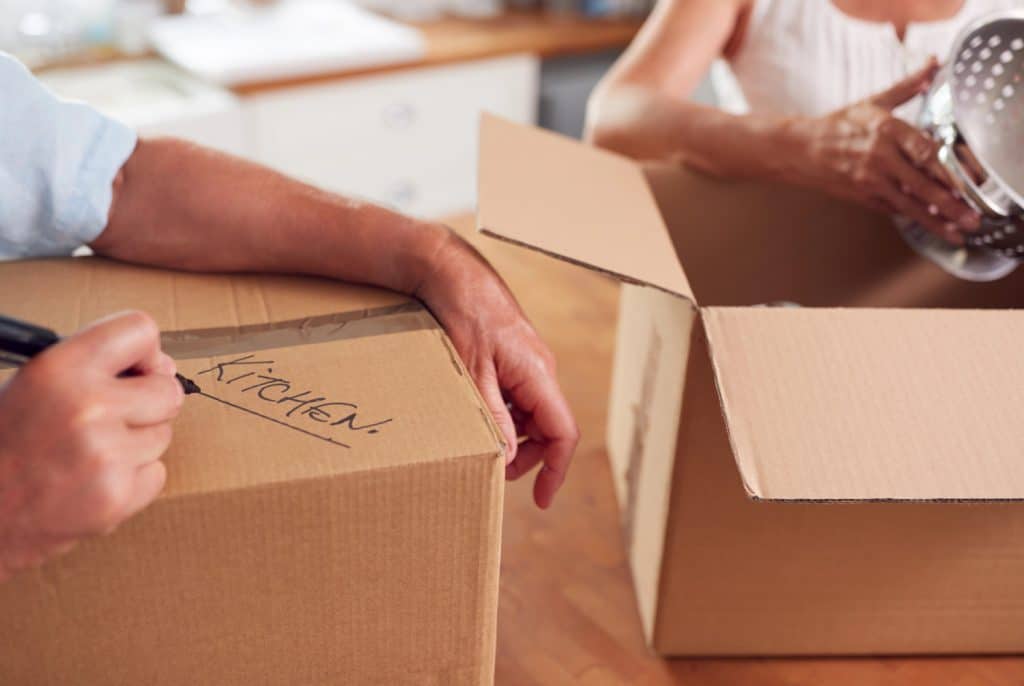 hands writing kitchen on a box next to hands packing a colander